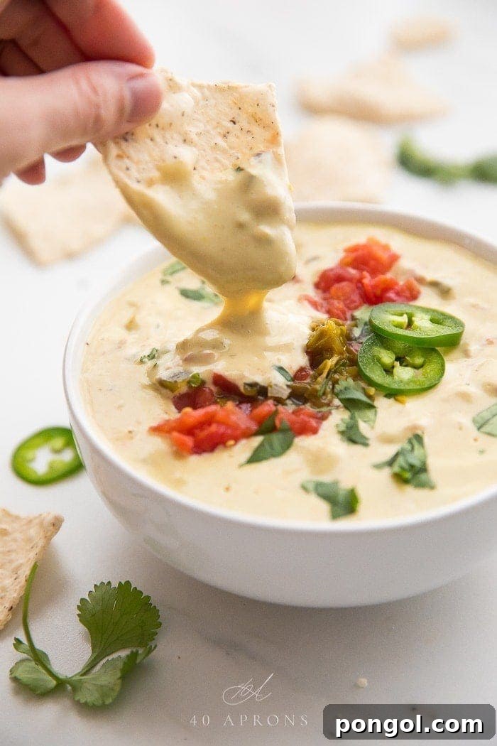 Hand dipping a tortilla chip into a bowl of vegan queso topped with tomatoes and jalapenos