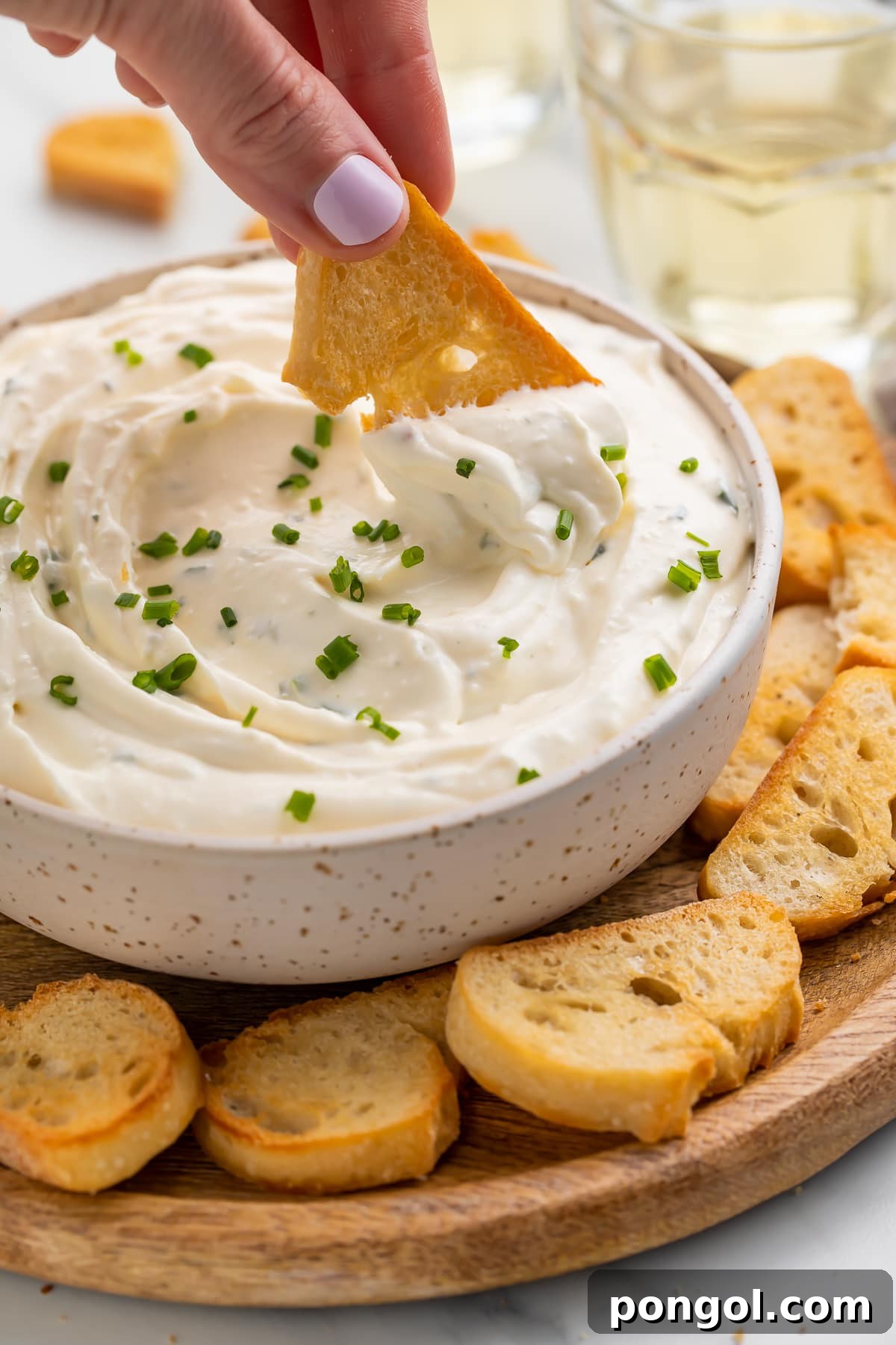Dreamy 3-Ingredient Boursin Dip 2 A hand scooping creamy Boursin dip with a crostini from a white bowl, showcasing the dip's smooth and inviting texture.