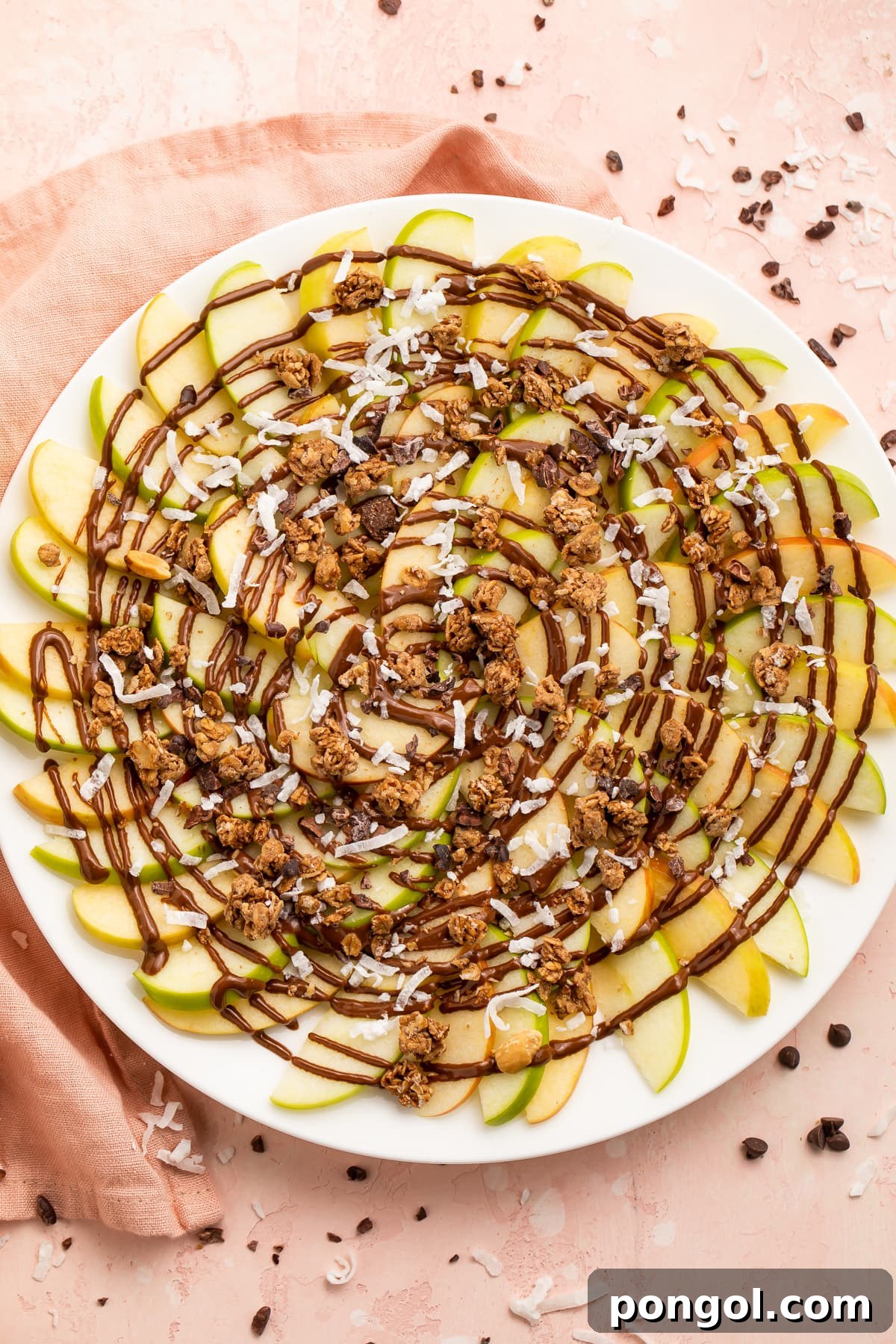 Smart Apple Stacks 3 Overhead, top-down view of a colorful plate of apple nachos featuring both green and red apple slices. They are generously coated with delicious swirls of chocolate syrup and sprinkled with crunchy granola, creating an inviting and healthy snack.