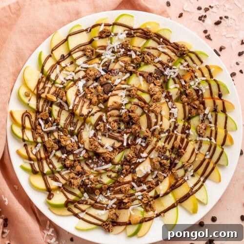 Overhead, top-down look at a plate of green and red apple slices topped with swirls of chocolate syrup and granola.