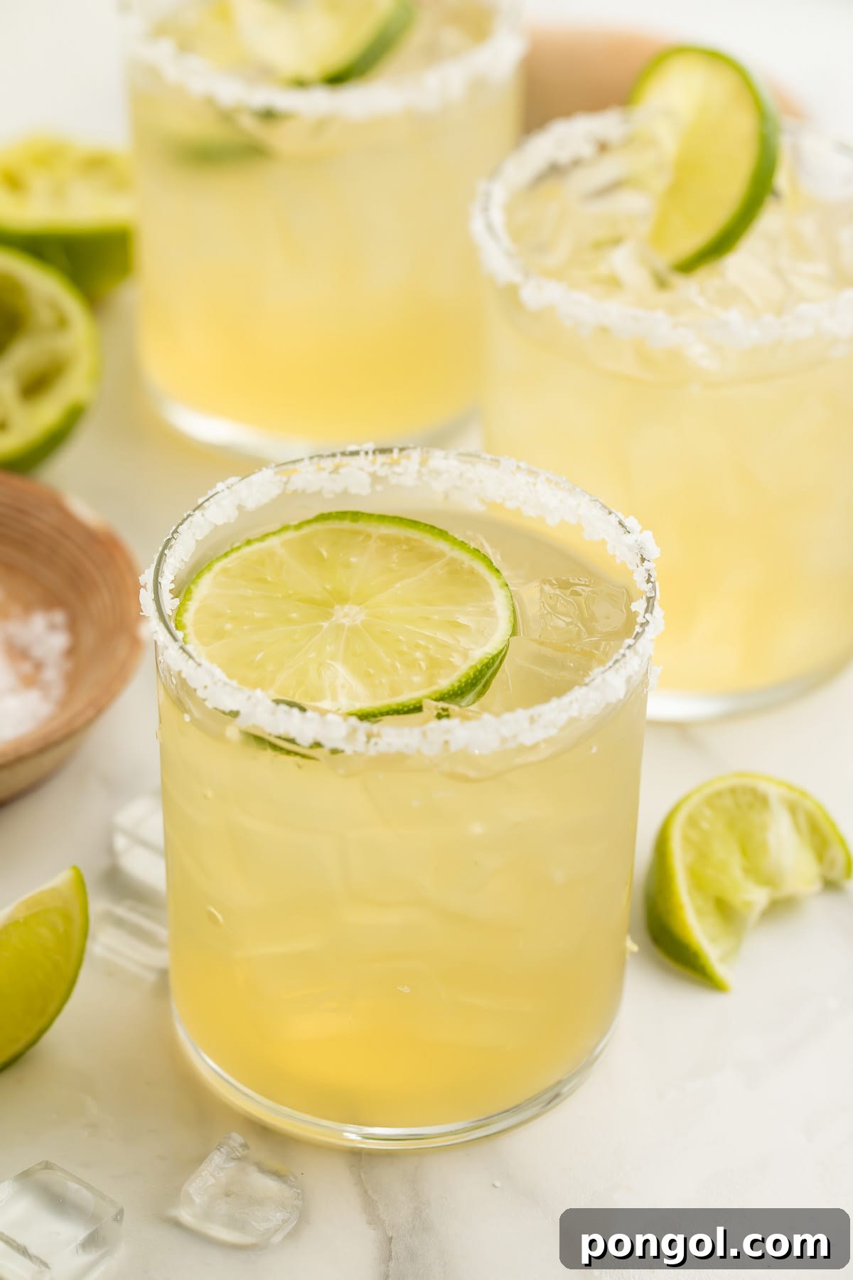 A salt-rimmed glass of virgin margarita garnished with a lime coin in front of 2 other glasses of margaritas.