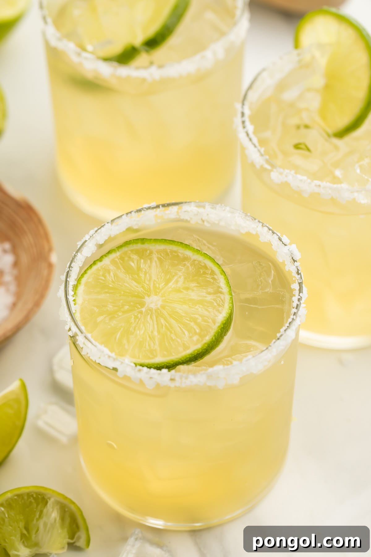 A salt-rimmed glass of virgin margarita garnished with a lime coin in front of 2 other glasses of margaritas.