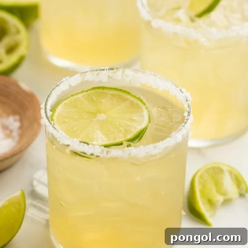 A salt-rimmed glass of virgin margarita garnished with a lime coin in front of 2 other glasses of margaritas.