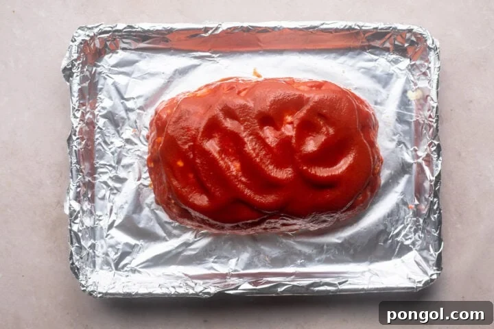 Hearty Gluten-Free Meatloaf 6 Unbaked meatloaf topped with a bright red ketchup glaze on a baking sheet lined with aluminum foil.
