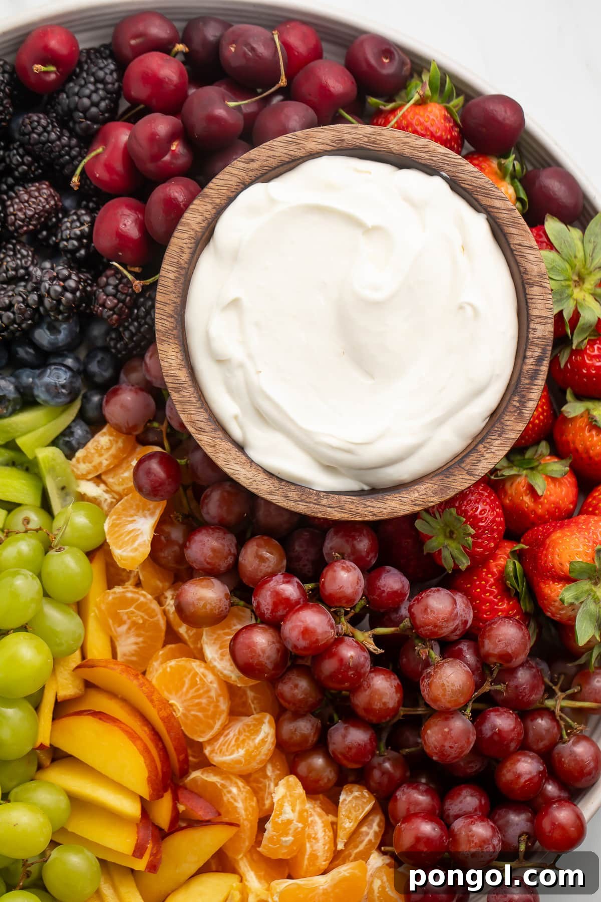 A small wooden bowl containing a white, whipped cream cheese dip on a platter surrounded by a rainbow of fruit.