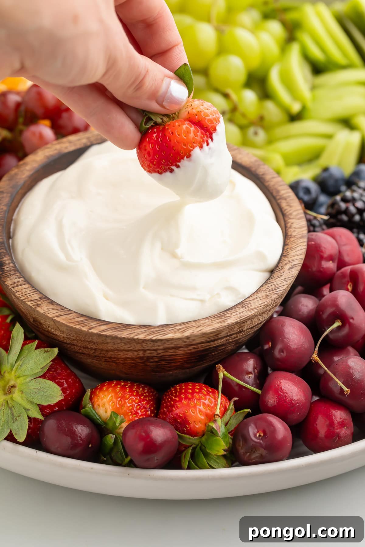 A white woman's hand holding a fresh strawberry as the woman dips it into a bowl of whipped cream cheese.