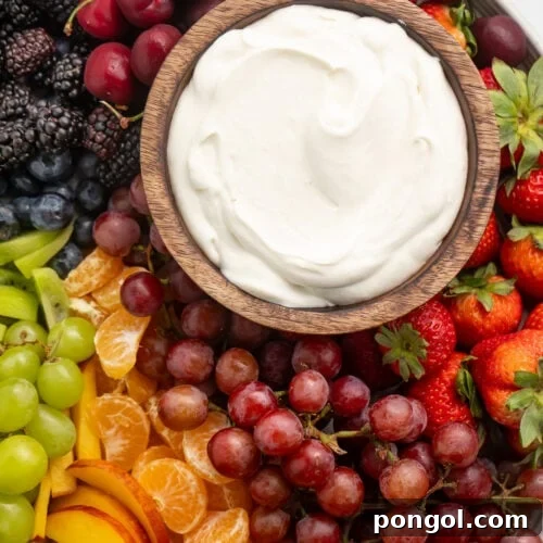 A small wooden bowl containing a white, whipped cream cheese dip on a platter surrounded by a rainbow of fruit.