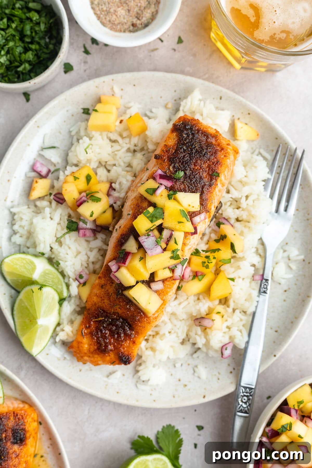 Jerk salmon topped with mango and red onion salsa on a plate with white rice and lime wedges.