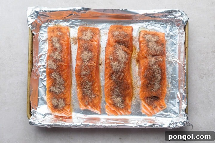 Four salmon fillets on a foil-lined baking sheet, topped with neutral oil and jerk seasoning.