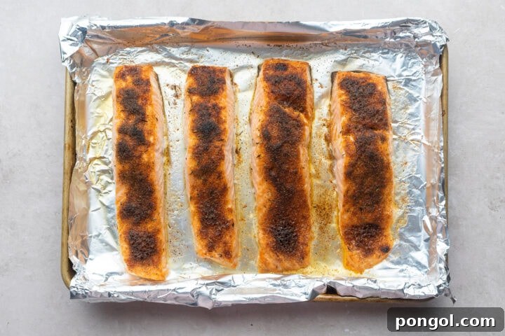 Four salmon fillets on a foil-lined baking sheet, topped with neutral oil and jerk seasoning.