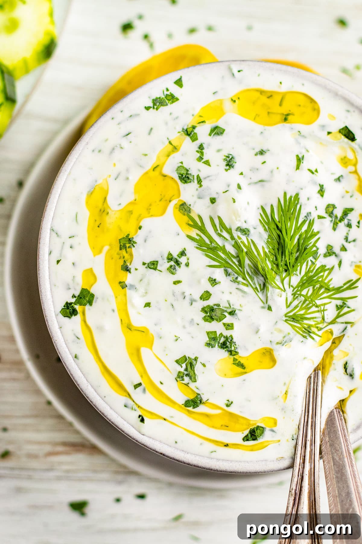 Close-up of a white bowl containing white Whole30 tzatziki dotted with bright green fresh herbs and topped with a drizzle of rich yellow olive oil.
