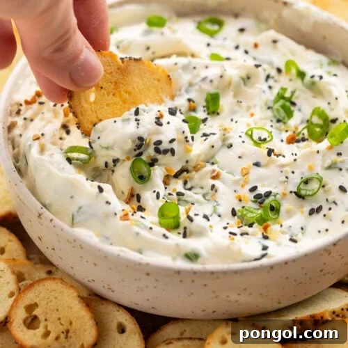 A white woman's hand dipping a bagel chip into a bowl of Everything Bagel dip surrounded by other bagel chips.
