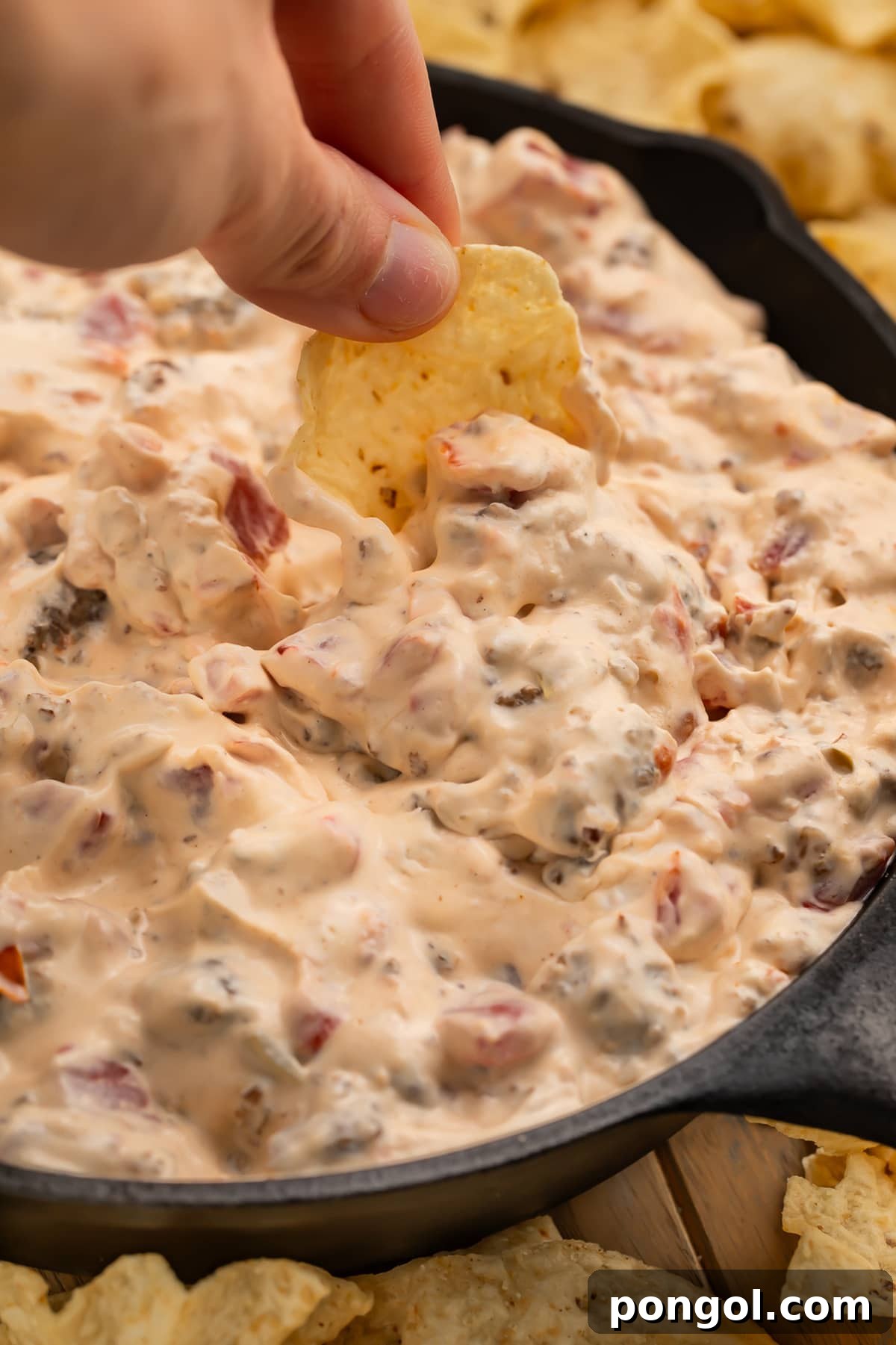 A white woman's hand dipping a scoop-shaped tortilla chip into a skillet full of cream cheese sausage dip.
