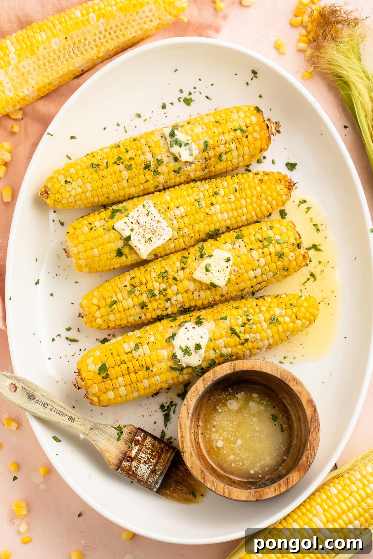 4 ears of air fryer corn on the cob arranged on a white oval platter next to a ramekin of melted butter.