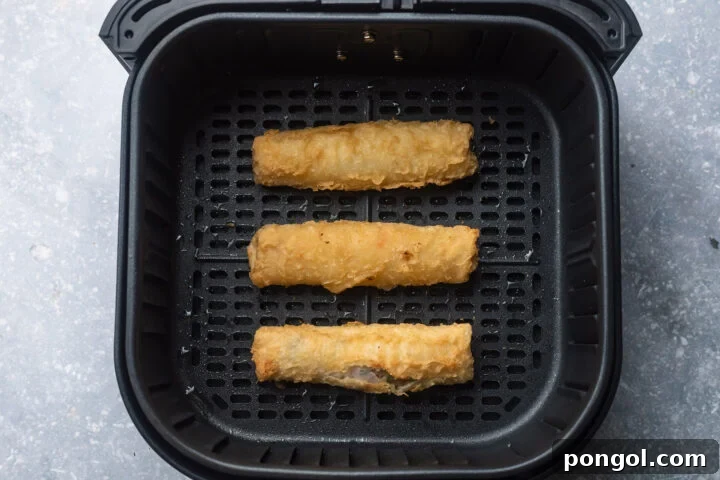 Three frozen egg rolls in a black air fryer basket.