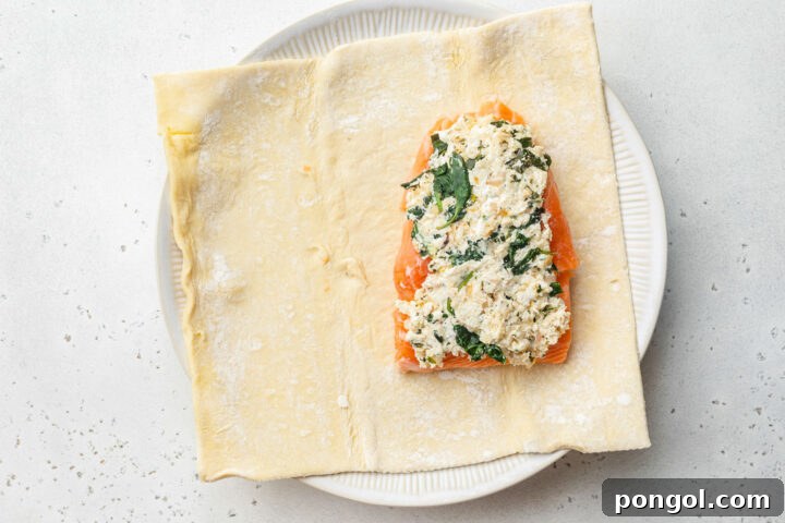 A sheet of puff pastry topped with a salmon fillet and cream cheese spinach filling on a white plate.