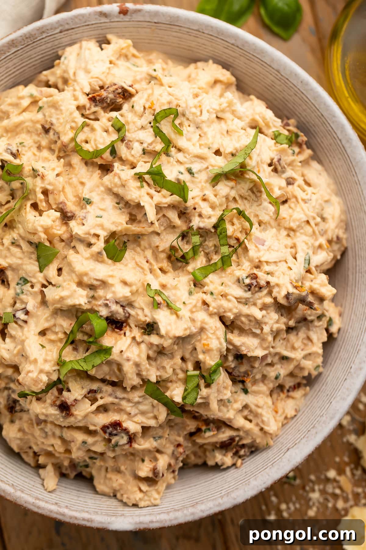 A close-up look at a large neutral bowl holding marry me chicken salad, with sun dried tomatoes and fresh basil, garnished with fresh basil leaves.