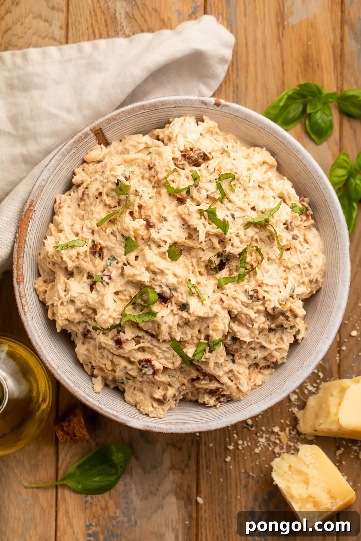 A zoomed-out look of a bowl of marry me chicken salad on a stained wood tabletop with a neutral cloth napkin, ready to be served.