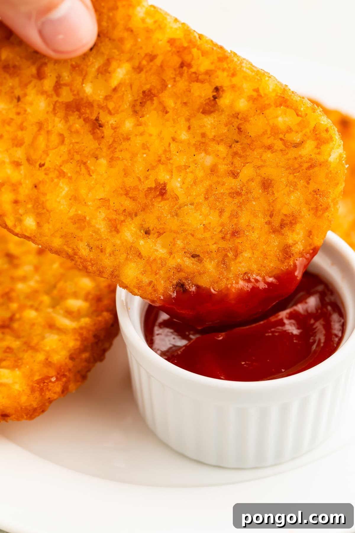 One crispy, air-fried hash brown patty being dipped into a small ramekin of red ketchup.