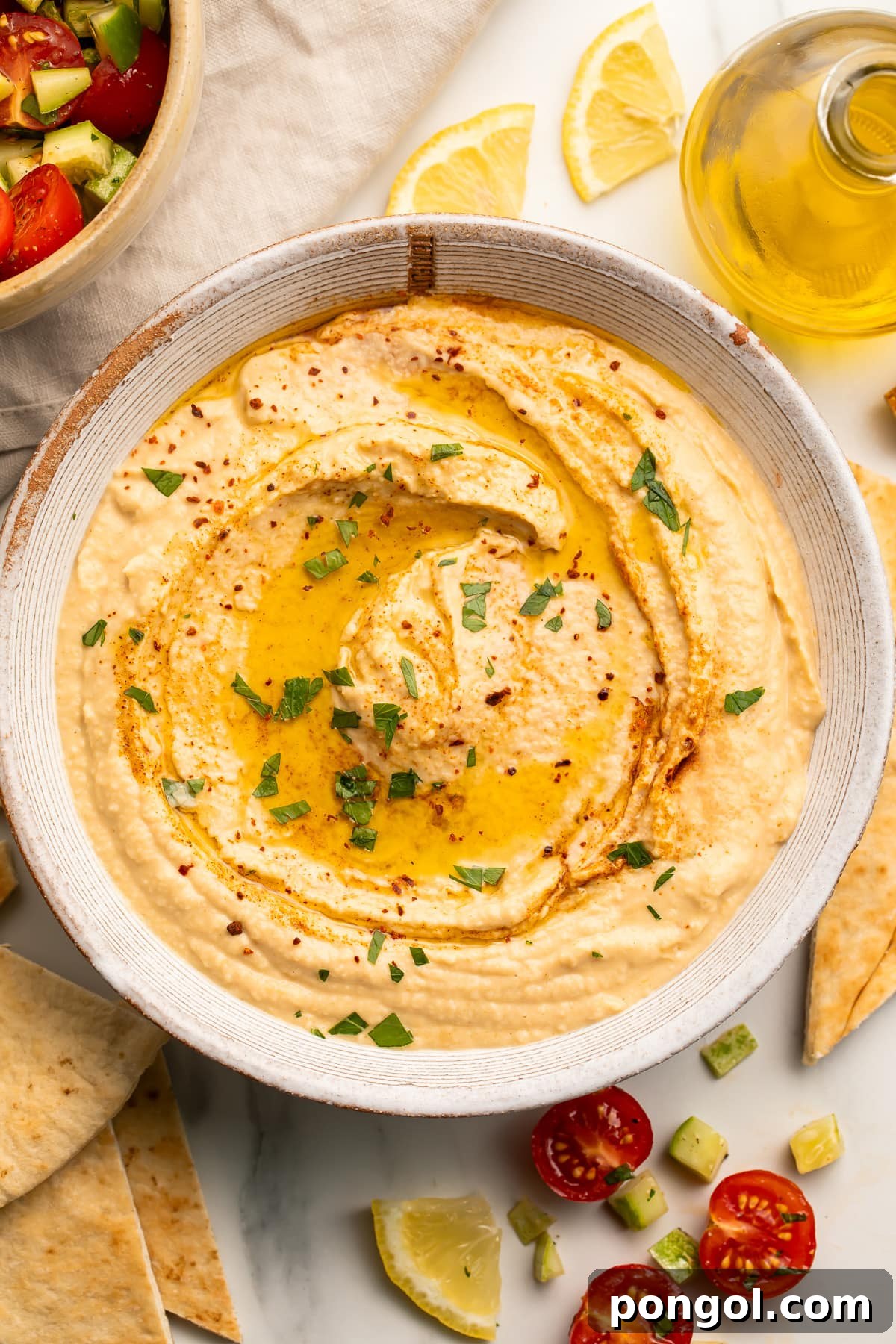 Creamy Tahini-Free Hummus 2 Overhead view of a white bowl holding creamy hummus made without tahini, swirled with olive oil and garnished with fresh herbs.