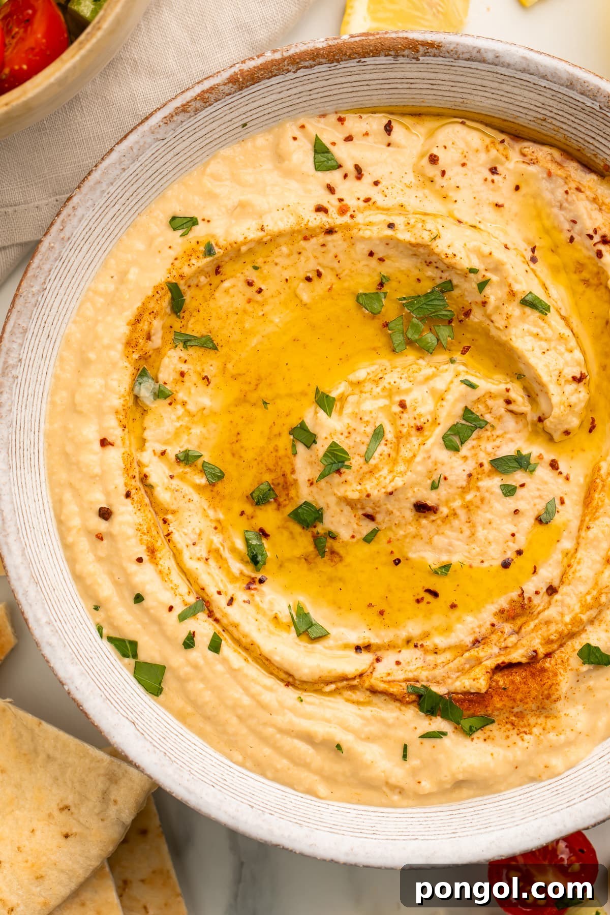 Creamy Tahini-Free Hummus 3 Close-up overhead view of a white bowl holding hummus made without tahini, swirled with olive oil and garnished with fresh herbs, emphasizing its creamy texture.