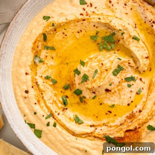 Close-up overhead view of a white bowl holding hummus made without tahini, swirled with olive oil and garnished with herbs.
