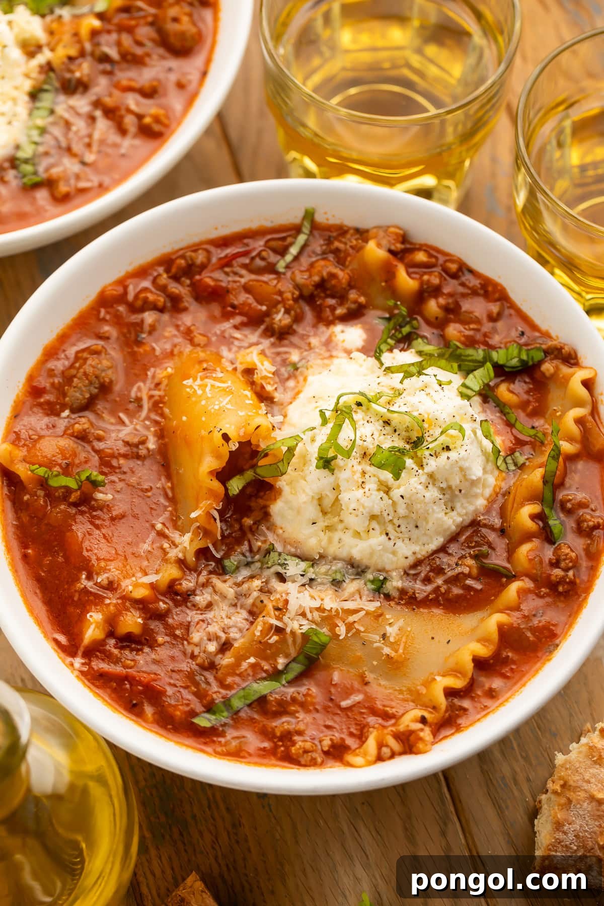 A bowl of lasagna soup on a wooden table next to a piece of bread and a glass of wine.