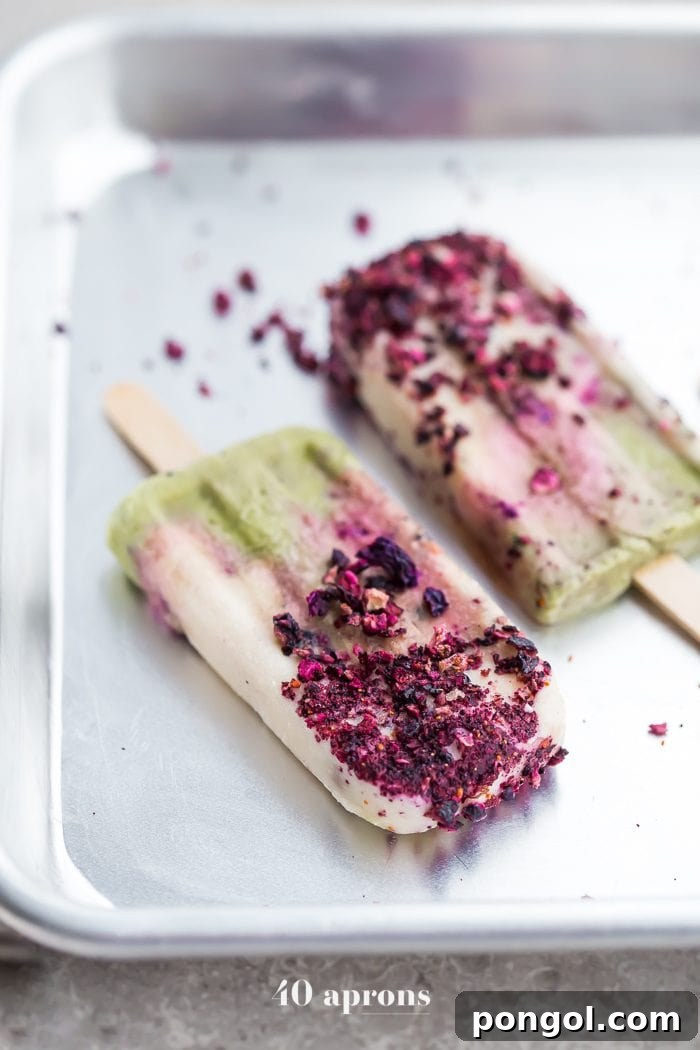 Hand holding a beautiful mermaid popsicle with distinct green, white, and purple layers, and a delicate topping of crushed freeze-dried blueberries.