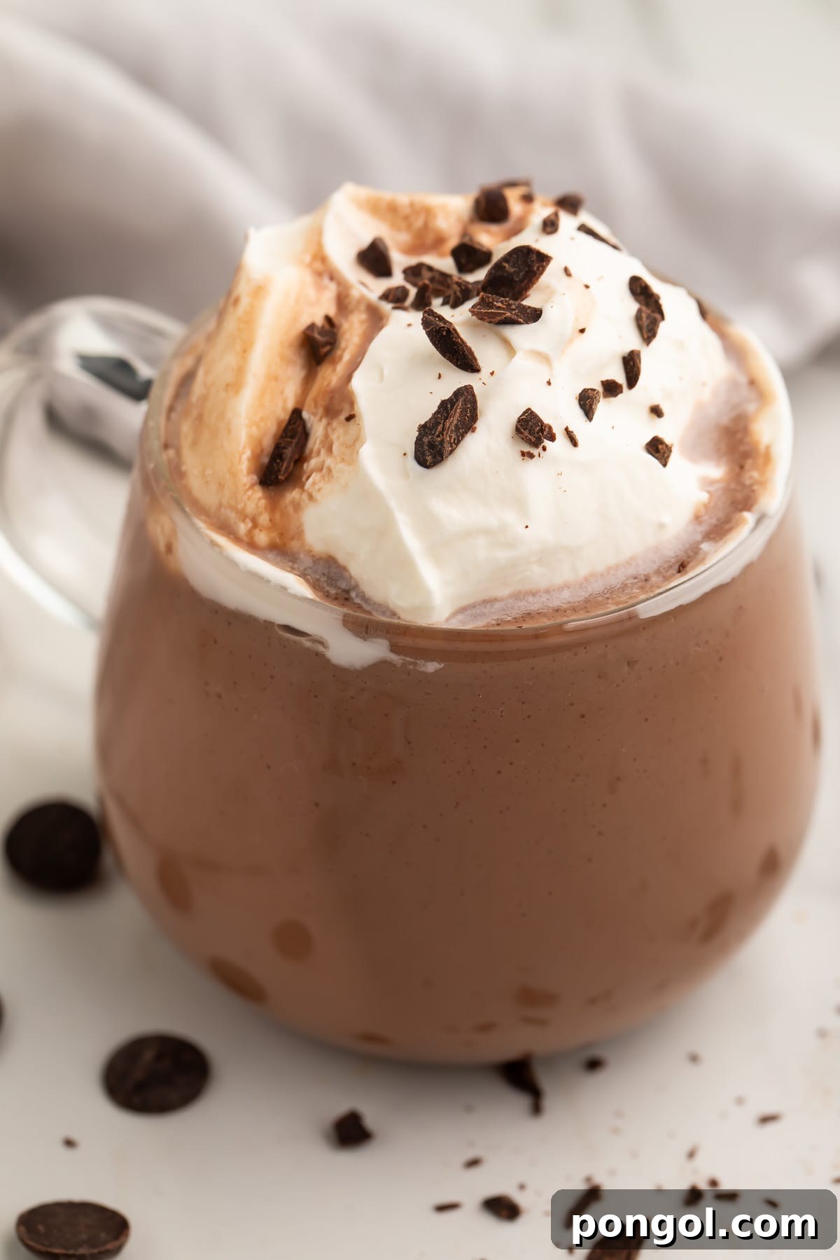 A glass mug filled with deep, rich paleo hot chocolate, generously topped with fluffy whipped coconut cream and delicate chocolate shavings. Perfect for winter warmth.