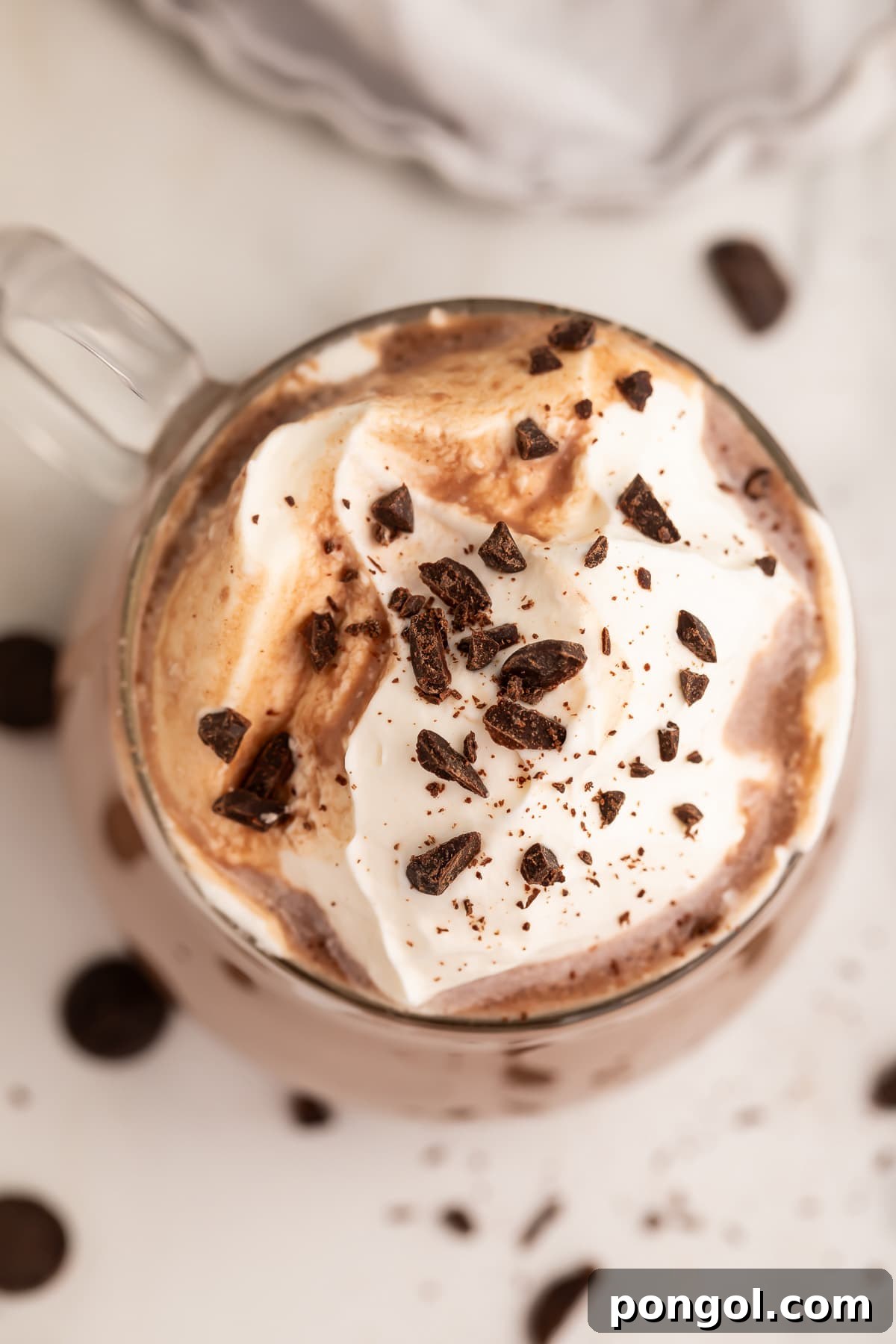 A top-down view of a clear glass coffee mug holding a rich, dark paleo hot chocolate, crowned with a dollop of whipped coconut cream and sprinkled with chocolate shavings.