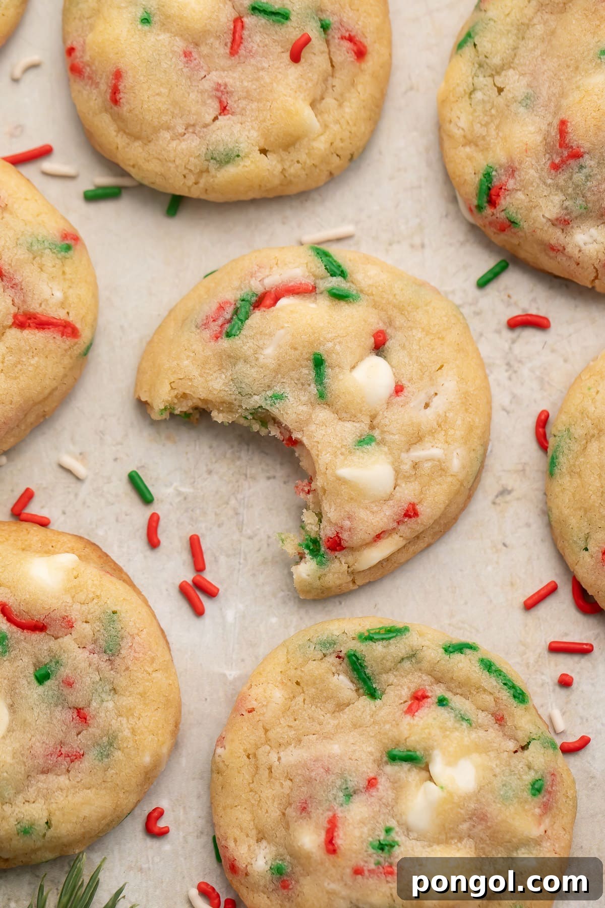 Festive Funfetti Cookies 3 A close-up shot of several Christmas confetti cookies on a sheet of parchment paper, adorned with vibrant red, white, and green sprinkles. One cookie in the center has a bite removed, revealing its soft, chewy interior.