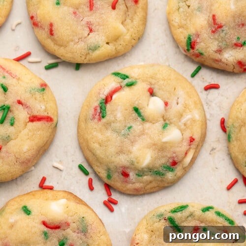 Rows of Christmas confetti cookies with red, white, and green sprinkles on parchment paper, with sprigs of rosemary resembling Christmas tree branches at the corners of the image.