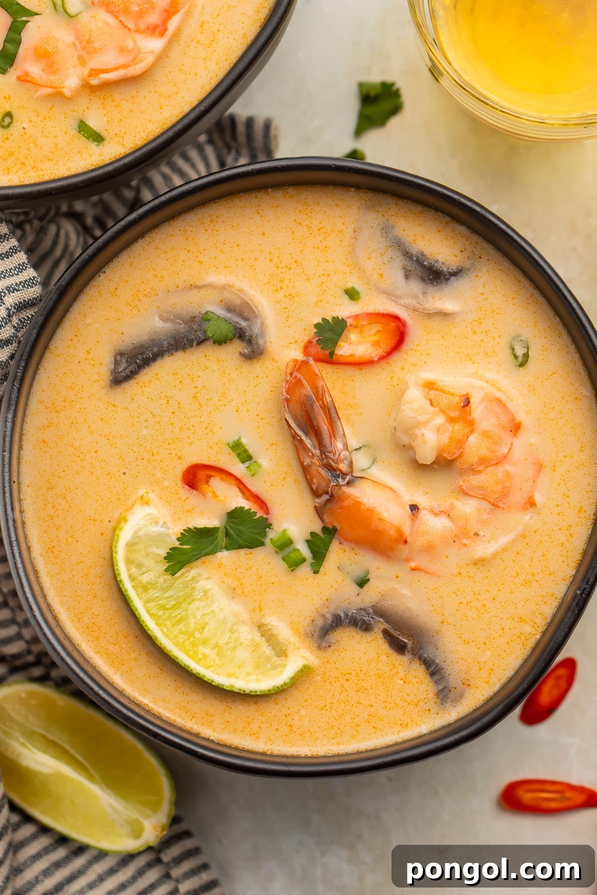 Tom Kha Goong: Creamy Thai Coconut Shrimp Delight 3 An overhead photo of a black soup bowl holding tom kha goong (Thai coconut shrimp soup) with lime wedges, shrimp, and aromatics floating in the broth.