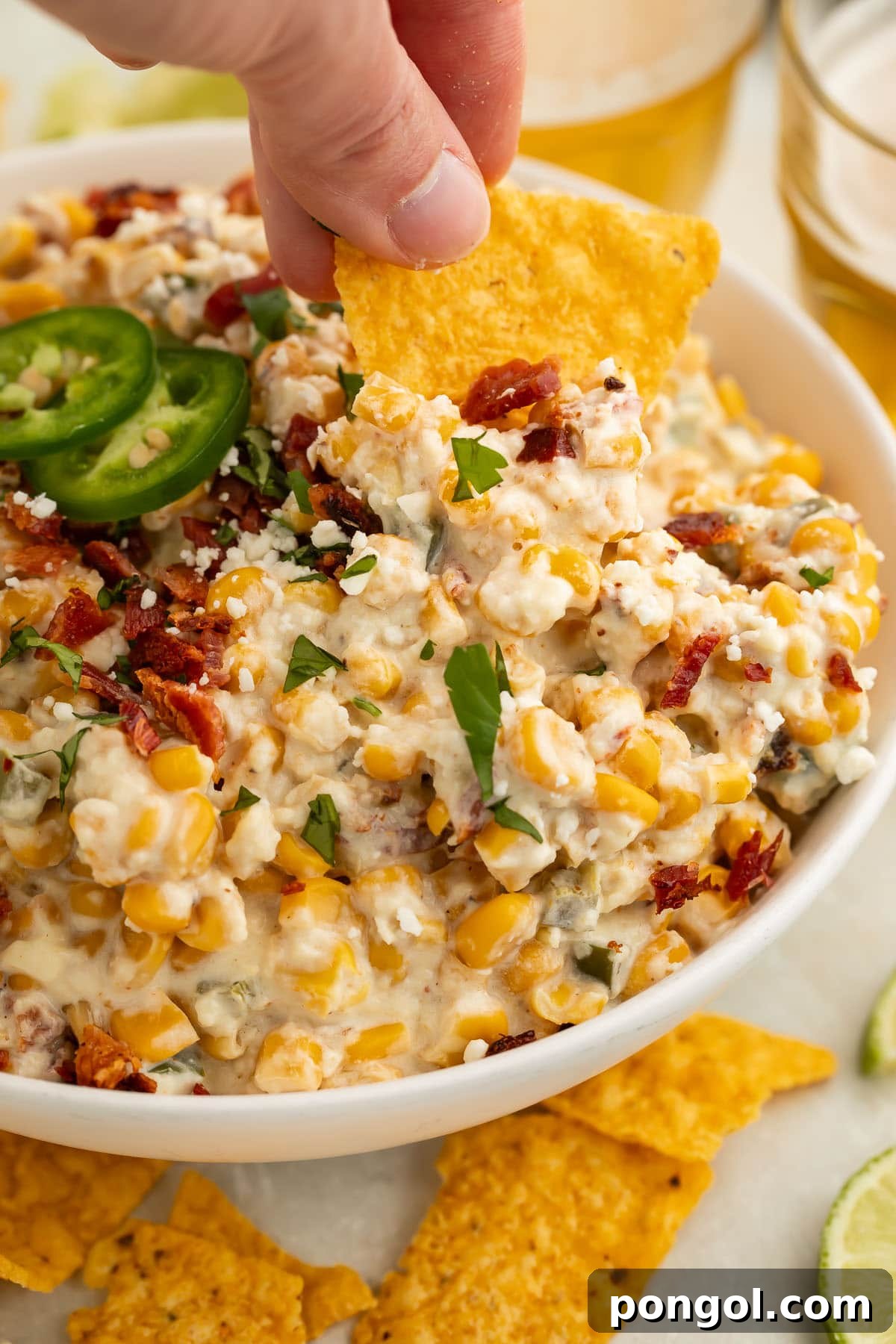 A hand holding a tortilla chip scooping creamy jalapeño corn dip from a large bowl, with crispy bacon and corn visible.
