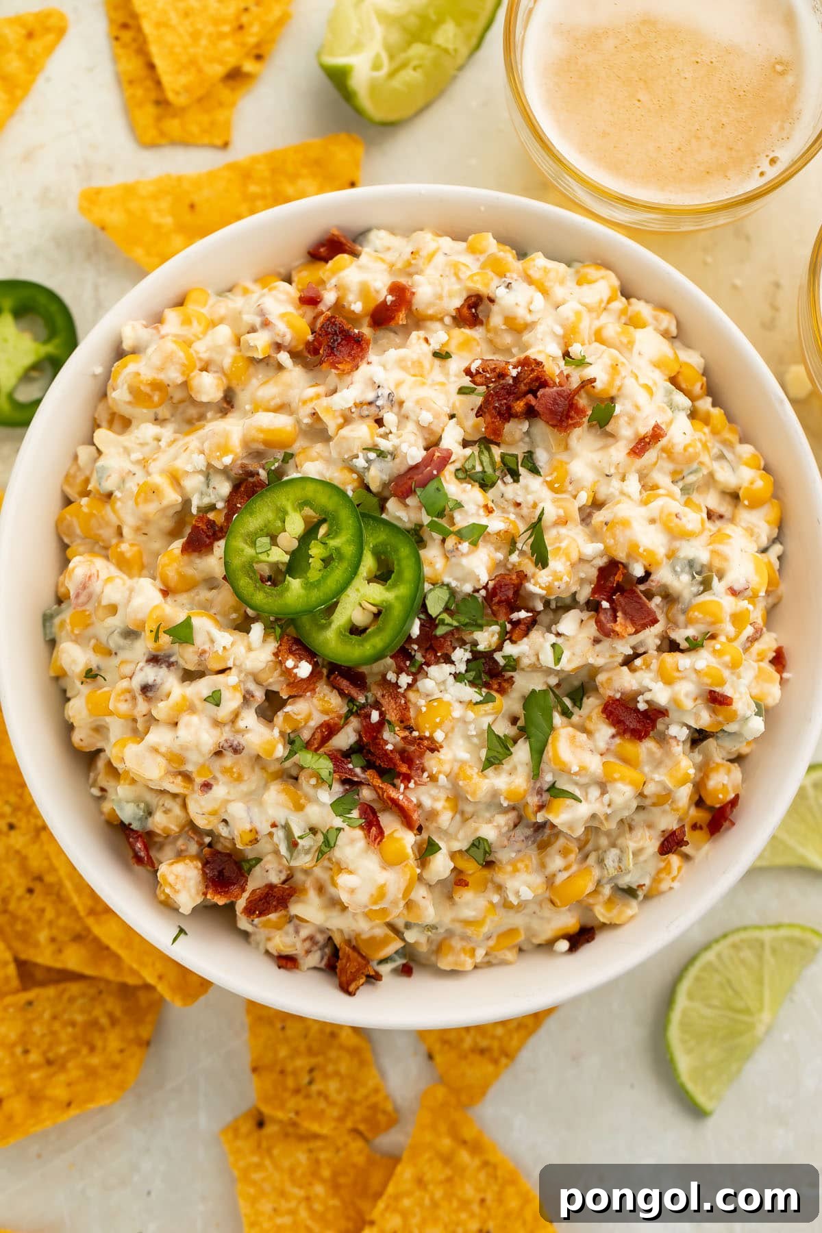 A top-down view of a large white bowl of jalapeño corn dip surrounded by golden tortilla chips on a rustic wooden table.