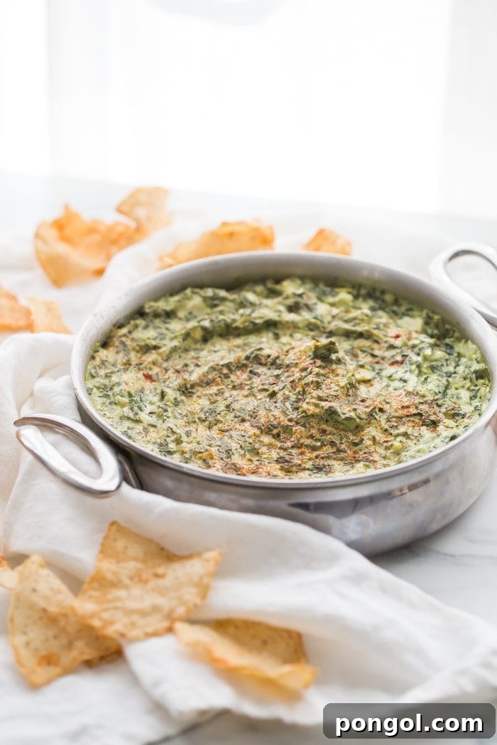 Creamy Paleo Vegan Spinach Artichoke Dip 8 A hand holding a paleo tortilla chip, scooping up creamy spinach artichoke dip