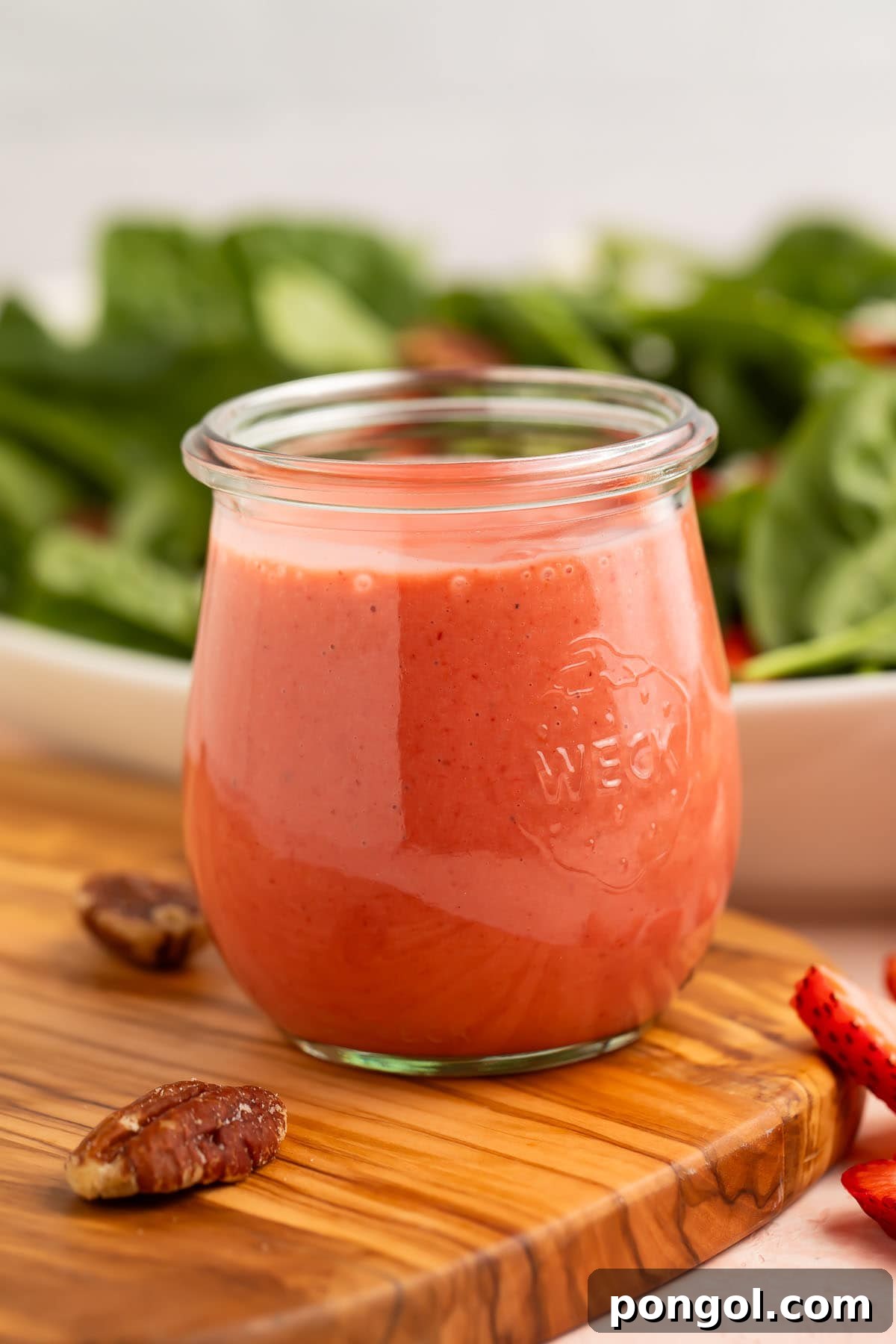 A jar of creamy pale red strawberry vinaigrette in front of a leafy spinach salad, highlighting its fresh and inviting appearance.