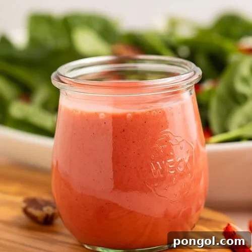 A jar of creamy pale red strawberry vinaigrette in front of a leafy spinach salad.