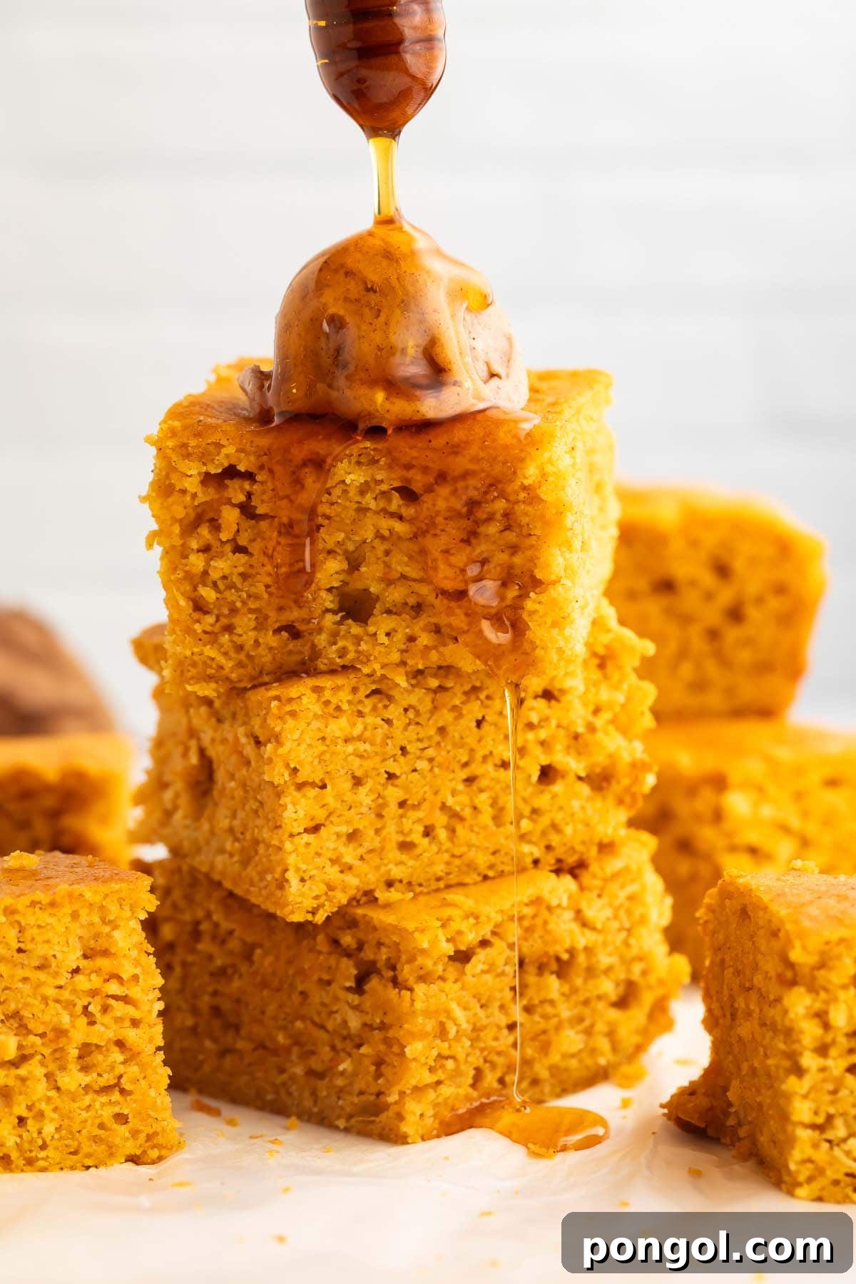 Golden sweet potato cornbread squares topped with dollops of cinnamon honey butter, with honey being drizzled over the stack, showcasing rich texture and inviting warmth.