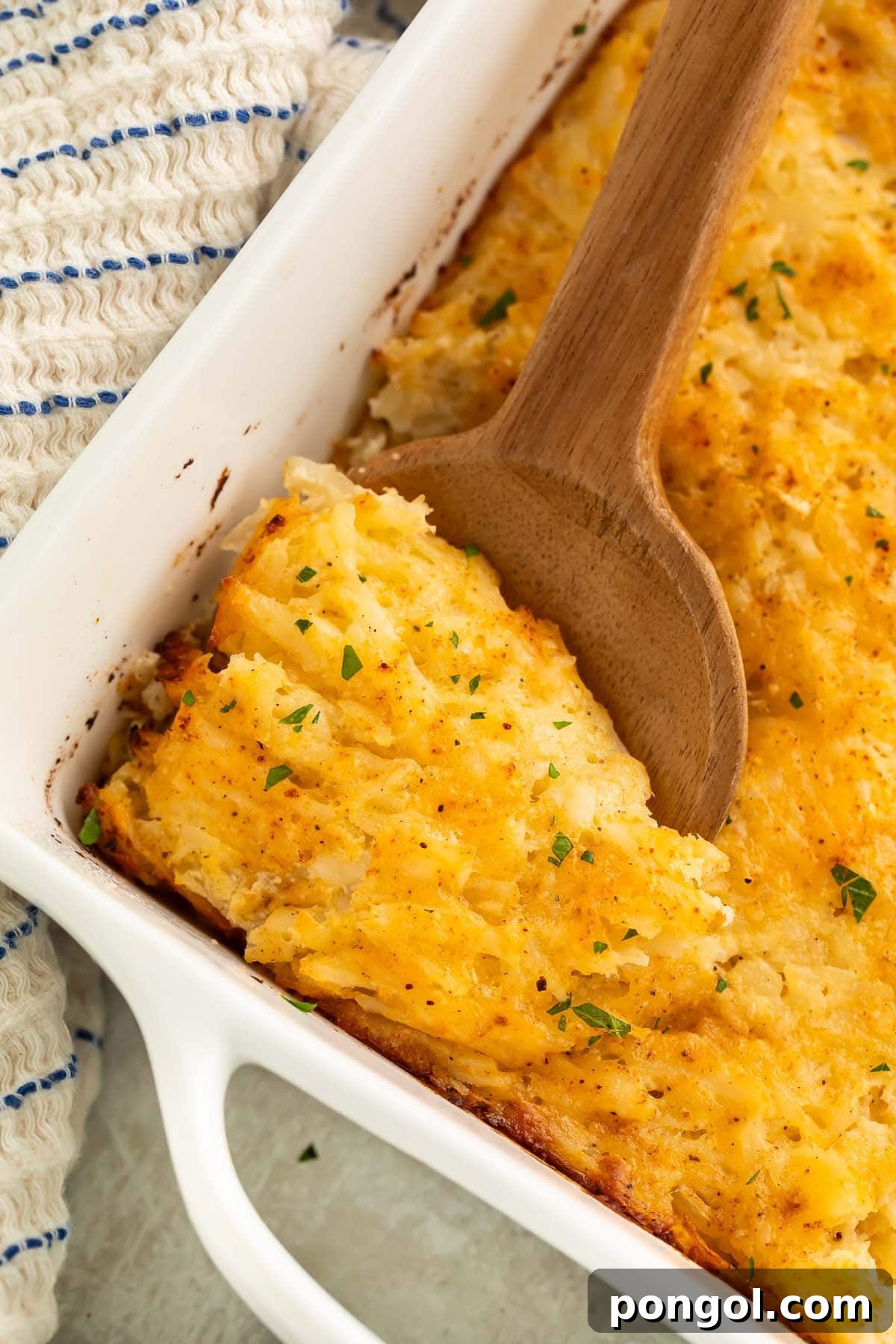A large wooden spoon resting in a square casserole dish holding a golden brown cheesy potato casserole, ready to be served.