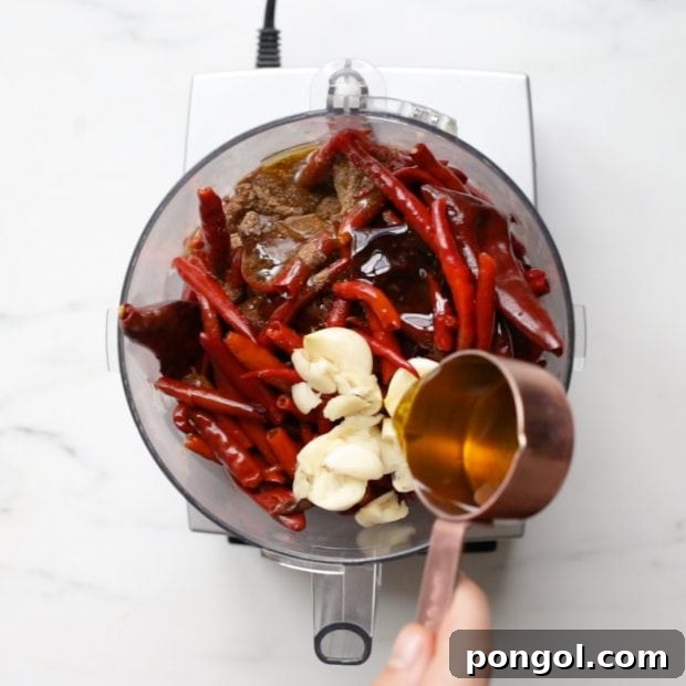 Premium olive oil being added to a food processor containing fresh and dried chilies and garlic, starting the blending process for harissa