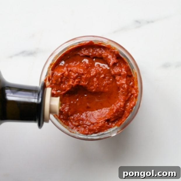 Olive oil being drizzled generously on top of a small glass jar filled with freshly made homemade harissa paste for preservation