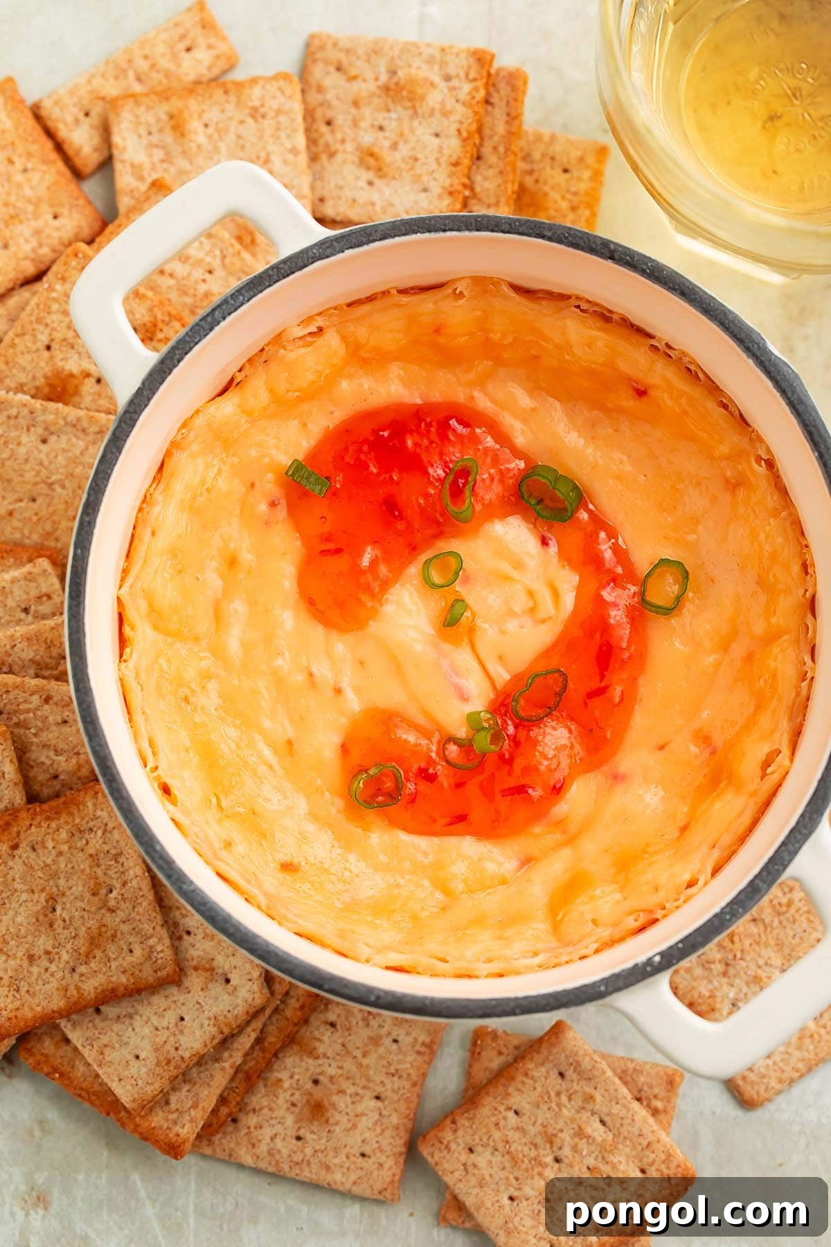 A high-sided bowl of creamy, baked cream cheese pepper jelly dip, artfully topped with a swirl of vibrant red pepper jelly and surrounded by perfectly arranged square wheat crackers, ready to be served.