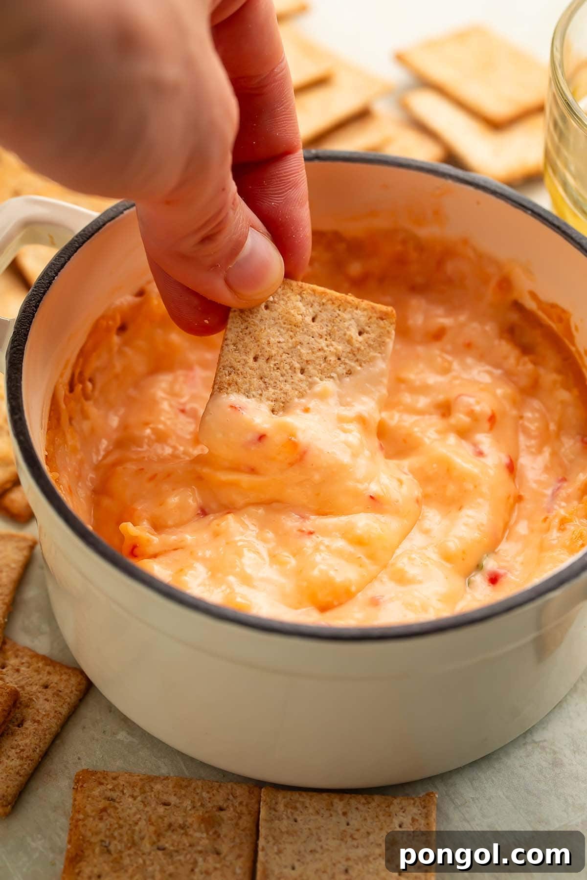 A woman's hand gracefully holding a square wheat cracker, poised to scoop into a generous bowl of creamy red pepper jelly dip, showcasing its inviting texture.