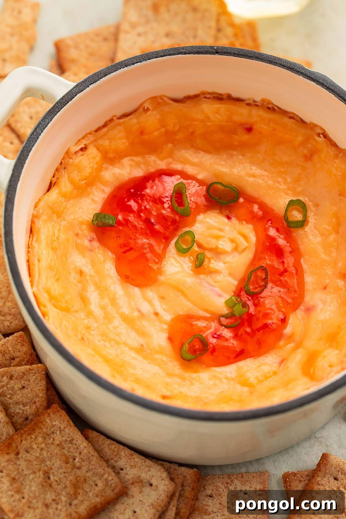 An overhead shot of a high-sided bowl filled with the baked cream cheese pepper jelly dip, adorned with a decorative swirl of red pepper jelly, surrounded by a variety of square wheat crackers, highlighting its presentation.