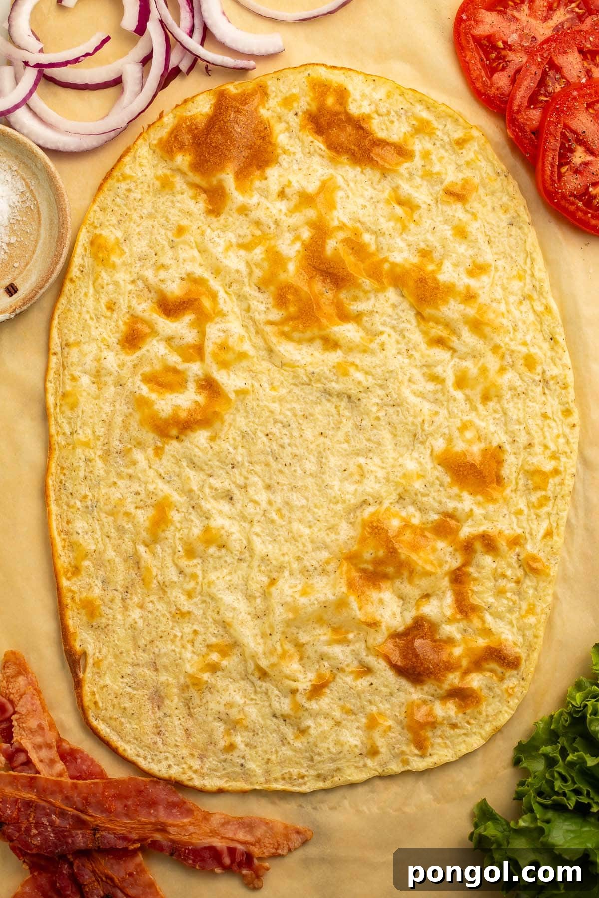 Creamy Cottage Cheese Flatbread Sensation 2 An unfolded cottage cheese flatbread on a sheet of parchment paper, beautifully golden-brown, surrounded by fresh sandwich ingredients like ham, bacon, lettuce, tomatoes, and onions, ready for assembly.