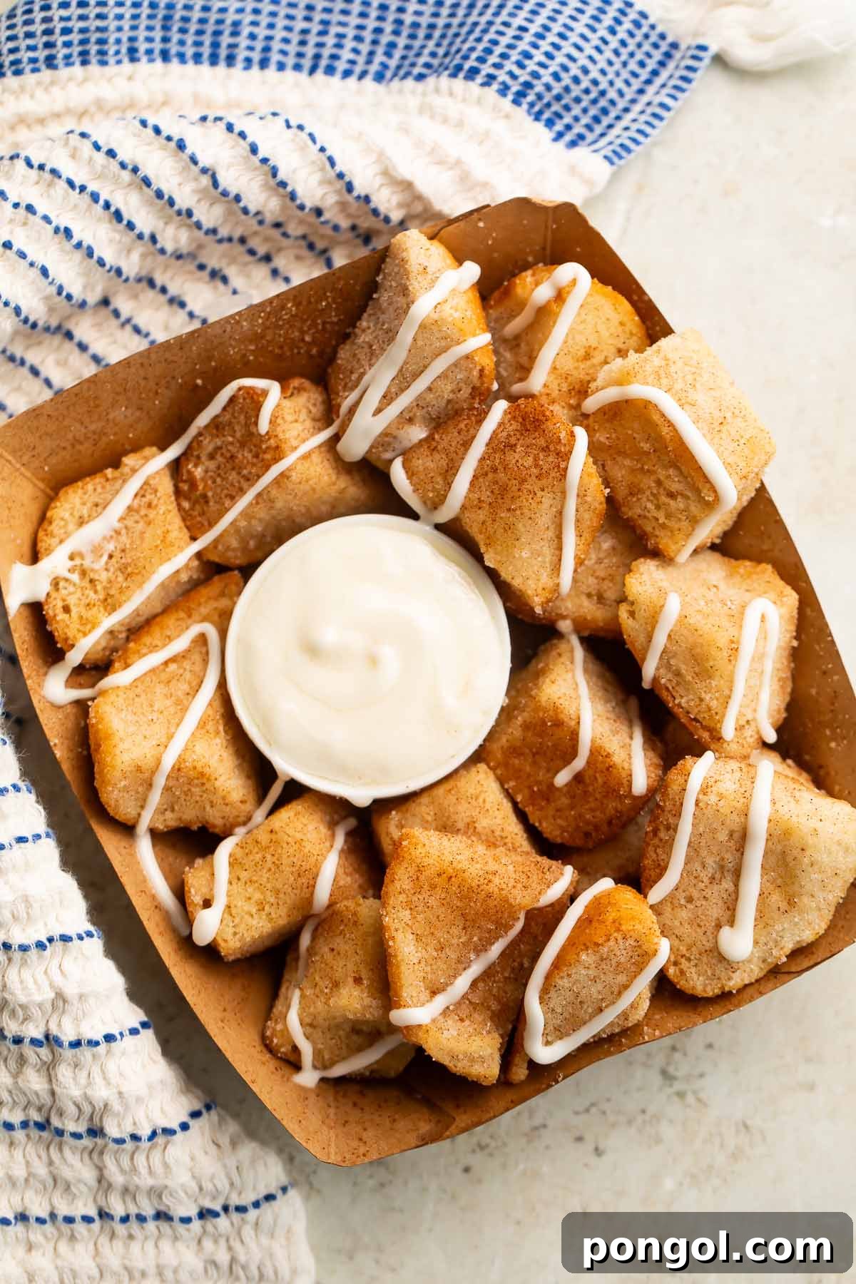 Overhead view of a cardboard tray filled with golden-brown keto cinnamon roll bites, ready to be dipped into a small ramekin of creamy white cream cheese icing.