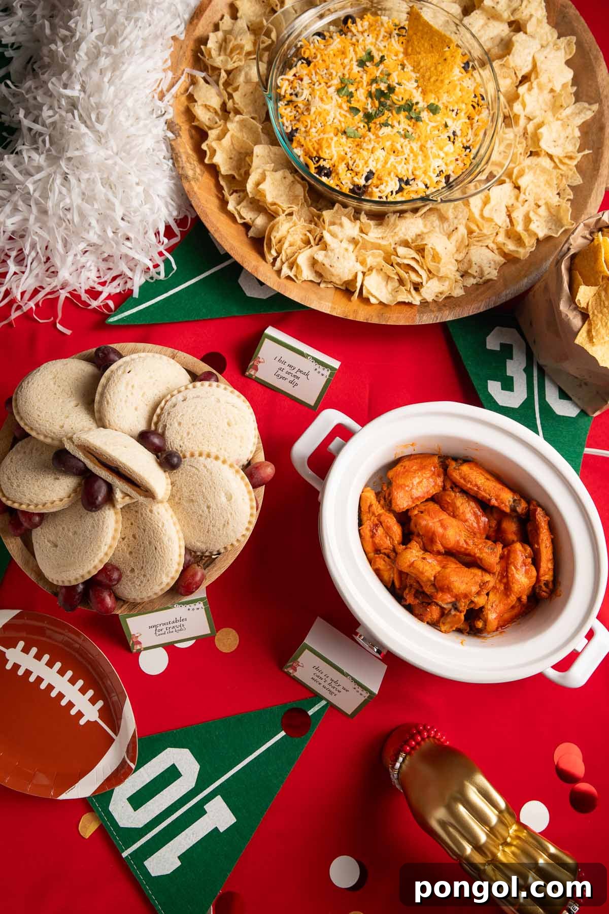 A table set for a Taylor Swift Super Bowl party, with football decorations, a red tablecloth, and gold heart hands placed among Crockpot chicken wings, Uncrustable sandwiches, and a 7 layer dip, showcasing a blend of Chiefs and Swiftie themes.