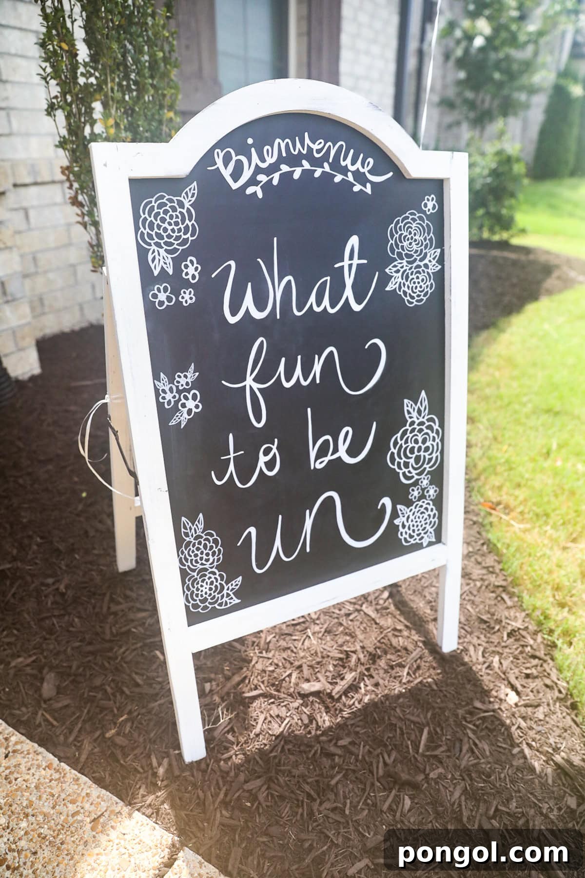 A free-standing white-framed chalkboard with the words "Bonjour! What fun to be un" written in cursive handwriting.