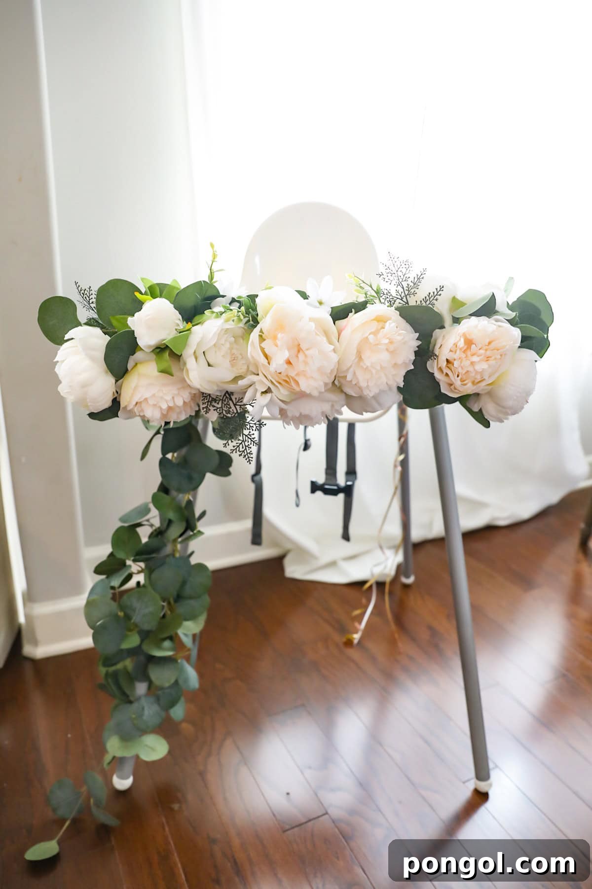 A white high chair decorated with faux white flowers and faux eucalyptus leaves.